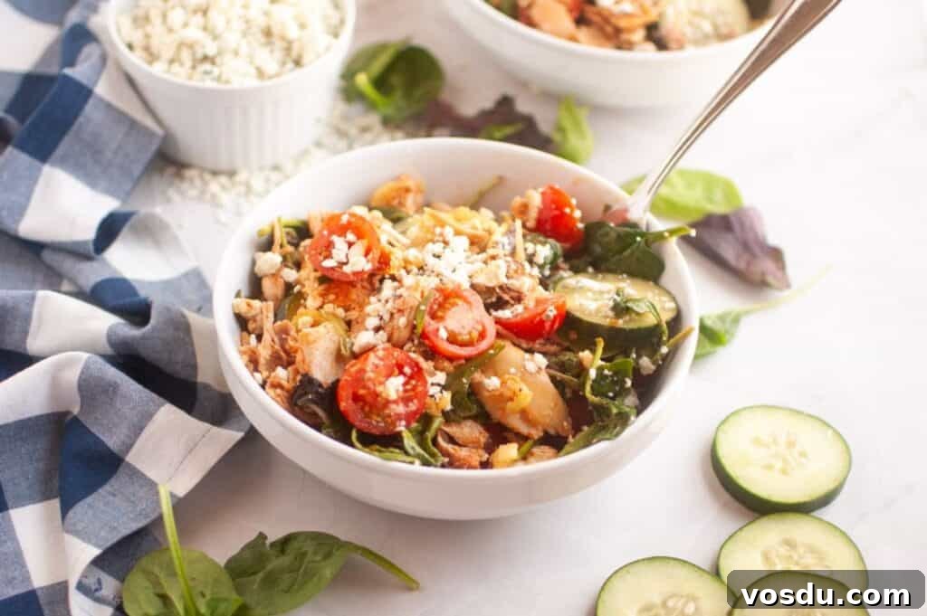 Chef Jenn’s Buffalo Chicken Salad Recipe for Flavorful Lunch 10 A bowl of salad with cherry tomatoes, cucumbers, spinach, chicken, and crumbled cheese, next to a blue checked napkin and sliced cucumber on a white surface.