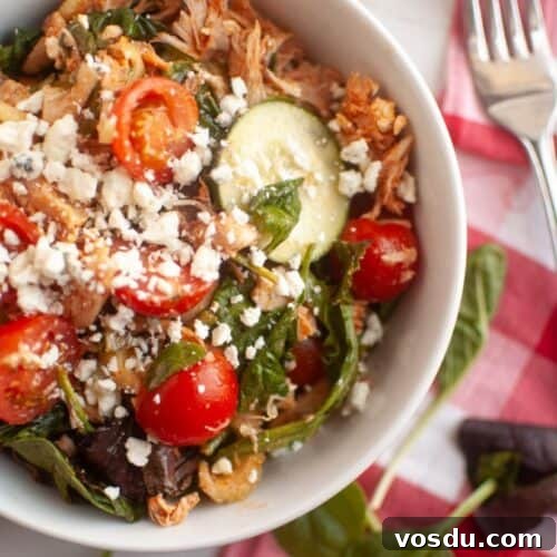 Chef Jenn’s Buffalo Chicken Salad Recipe for Flavorful Lunch 11 A bowl of salad with shredded meat, cherry tomatoes, spinach, cucumber slices, and crumbled cheese, placed next to a fork and a red-and-white checkered napkin.