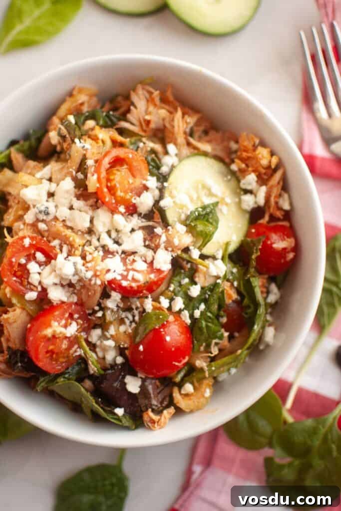 Chef Jenn’s Buffalo Chicken Salad Recipe for Flavorful Lunch 3 A white bowl filled with shredded chicken, cherry tomatoes, spinach, cucumber slices, and crumbled feta cheese, placed on a table with a fork and some spinach leaves nearby.