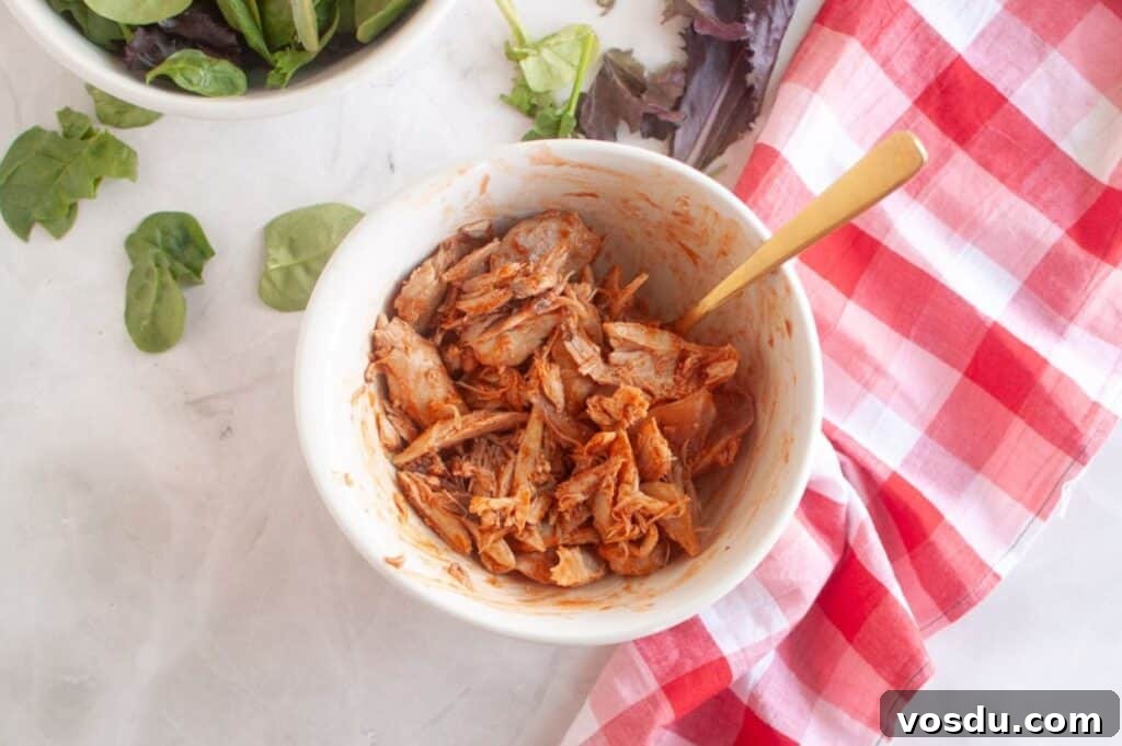 Chef Jenn’s Buffalo Chicken Salad Recipe for Flavorful Lunch 5 A bowl of shredded, seasoned jackfruit with a gold spoon, next to a bowl of mixed greens and a red checkered cloth on a white surface.