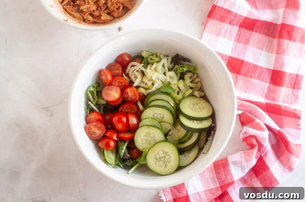 Chef Jenn’s Buffalo Chicken Salad Recipe for Flavorful Lunch 6 A white bowl filled with sliced cucumbers, cherry tomatoes, celery, and mixed greens sits next to a red and white checkered cloth on a light surface.