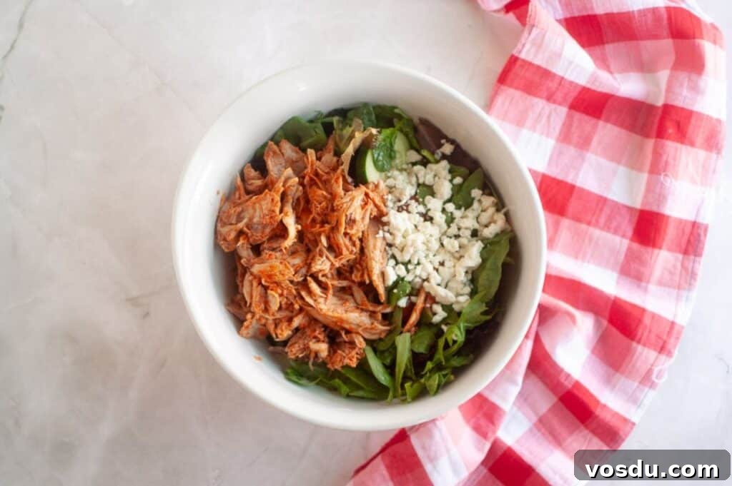 Chef Jenn’s Buffalo Chicken Salad Recipe for Flavorful Lunch 7 A white bowl containing shredded chicken, mixed greens, and crumbled cheese on a marble surface with a red and white checkered cloth beside it.