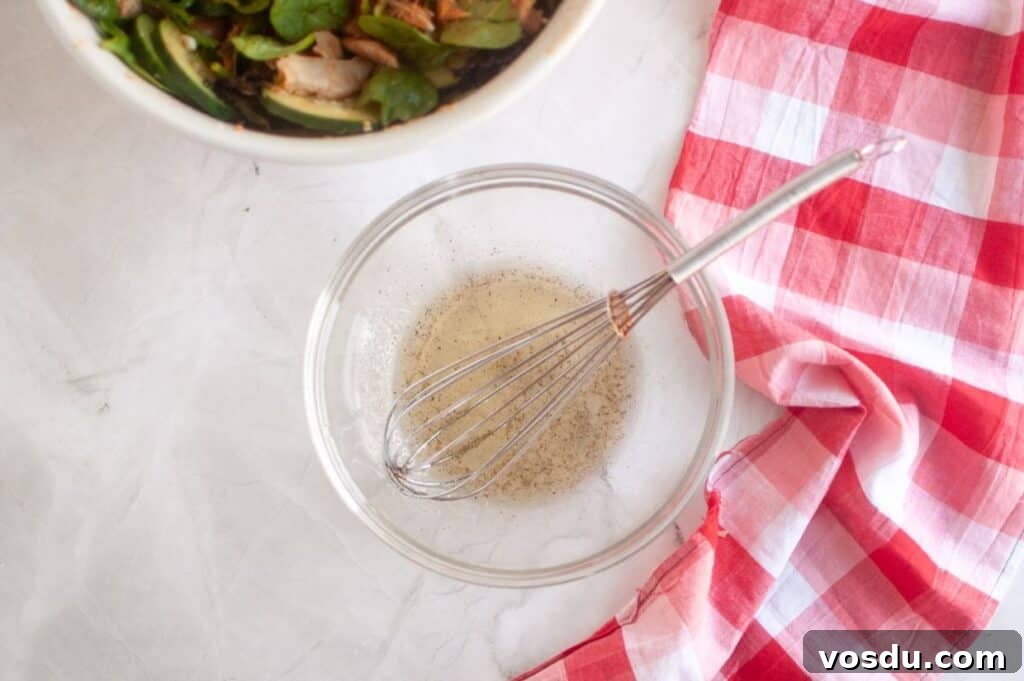 Chef Jenn’s Buffalo Chicken Salad Recipe for Flavorful Lunch 8 A glass bowl with whisked vinaigrette and a metal whisk beside a red and white checkered cloth; a salad bowl is partially visible in the corner.
