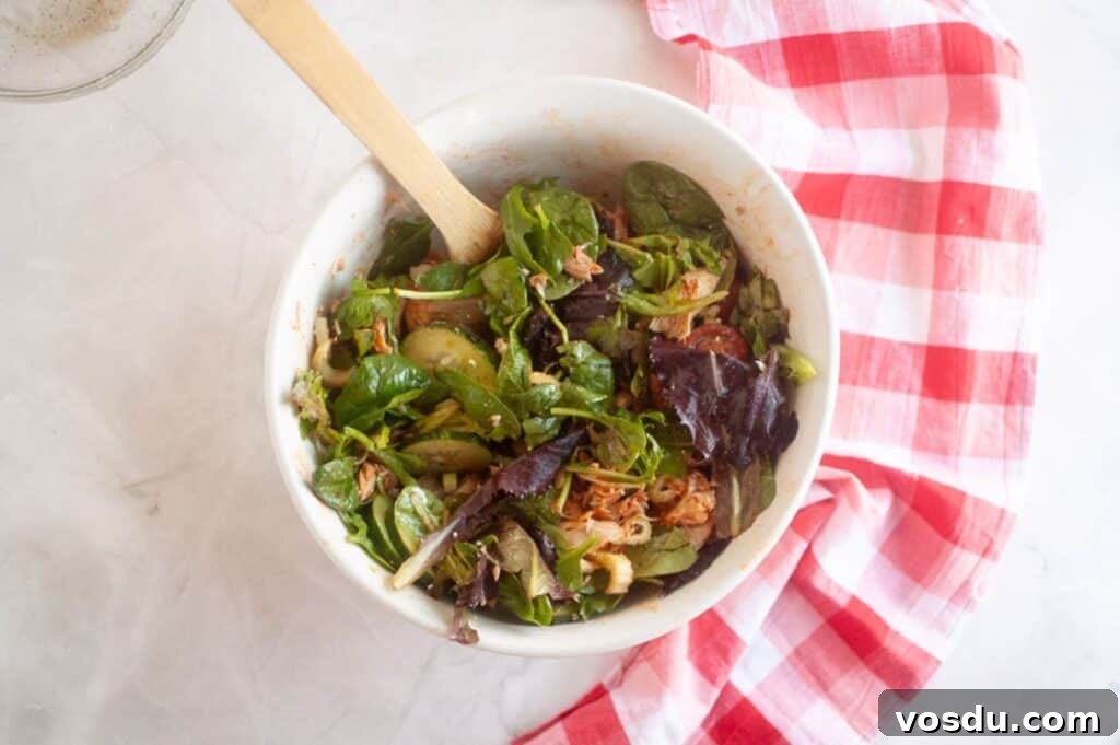 Chef Jenn’s Buffalo Chicken Salad Recipe for Flavorful Lunch 9 A white bowl filled with mixed greens, shredded meat, and sliced vegetables, with a wooden spoon, sits on a marble surface next to a red and white checkered cloth.