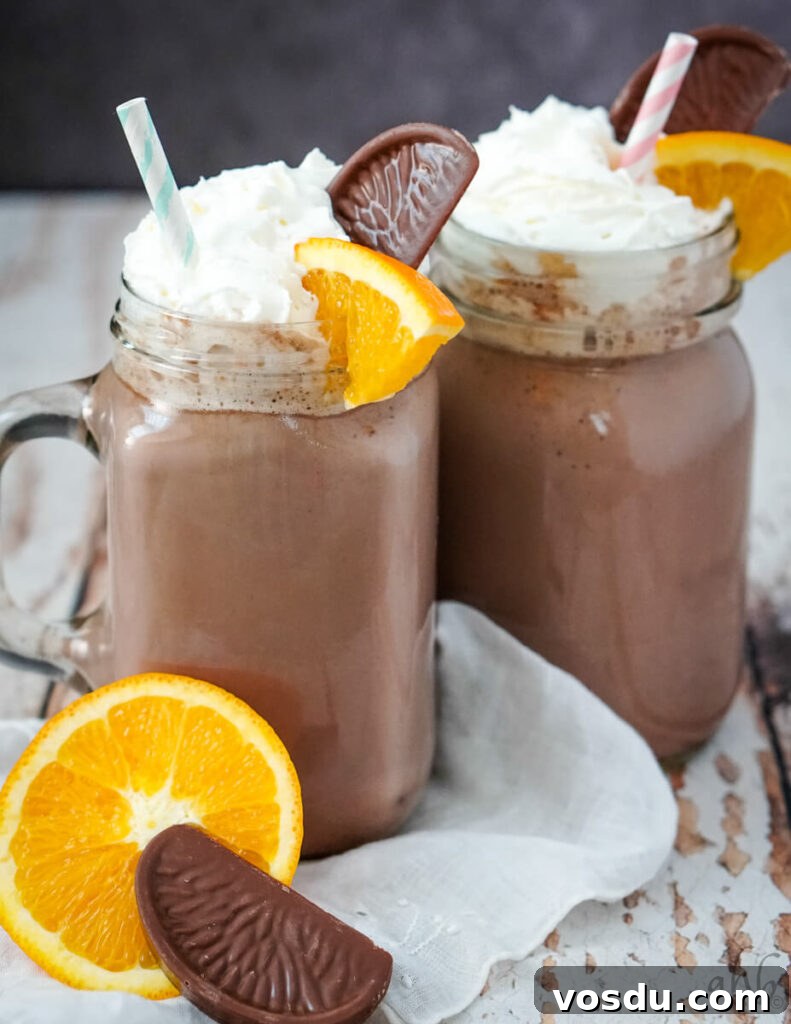Velvety Orange Hot Chocolate Recipe: Creamy Spiced Cocoa 4 Clear glass mugs full of orange hot chocolate with striped straws in them. An orange and a chocolate in front of the mugs.