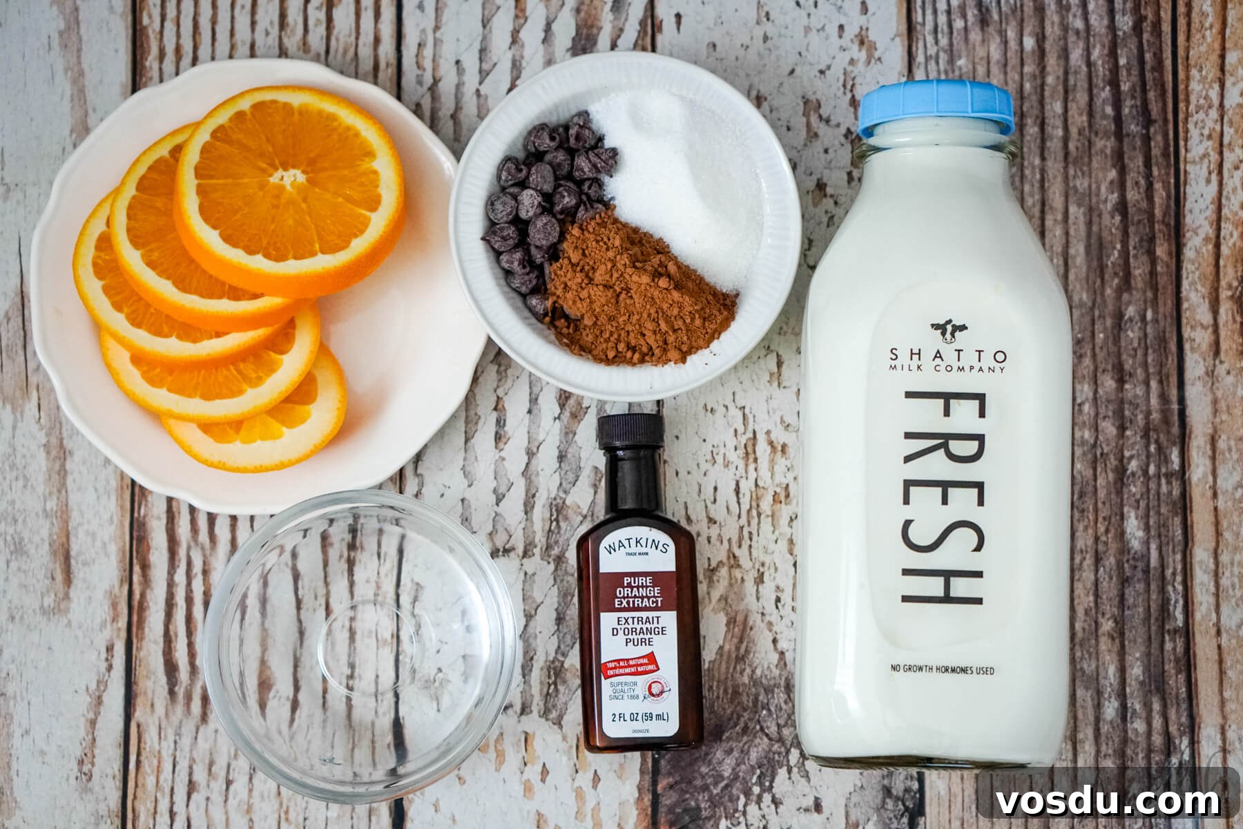 Velvety Orange Hot Chocolate Recipe: Creamy Spiced Cocoa 5 Slices of orange on a white plate, bowls of granulated sugar, cocoa powder, and semi-sweet chocolate chips in a white bowl, a bowl of water, a bottle of orange extract, and a bottle of milk.