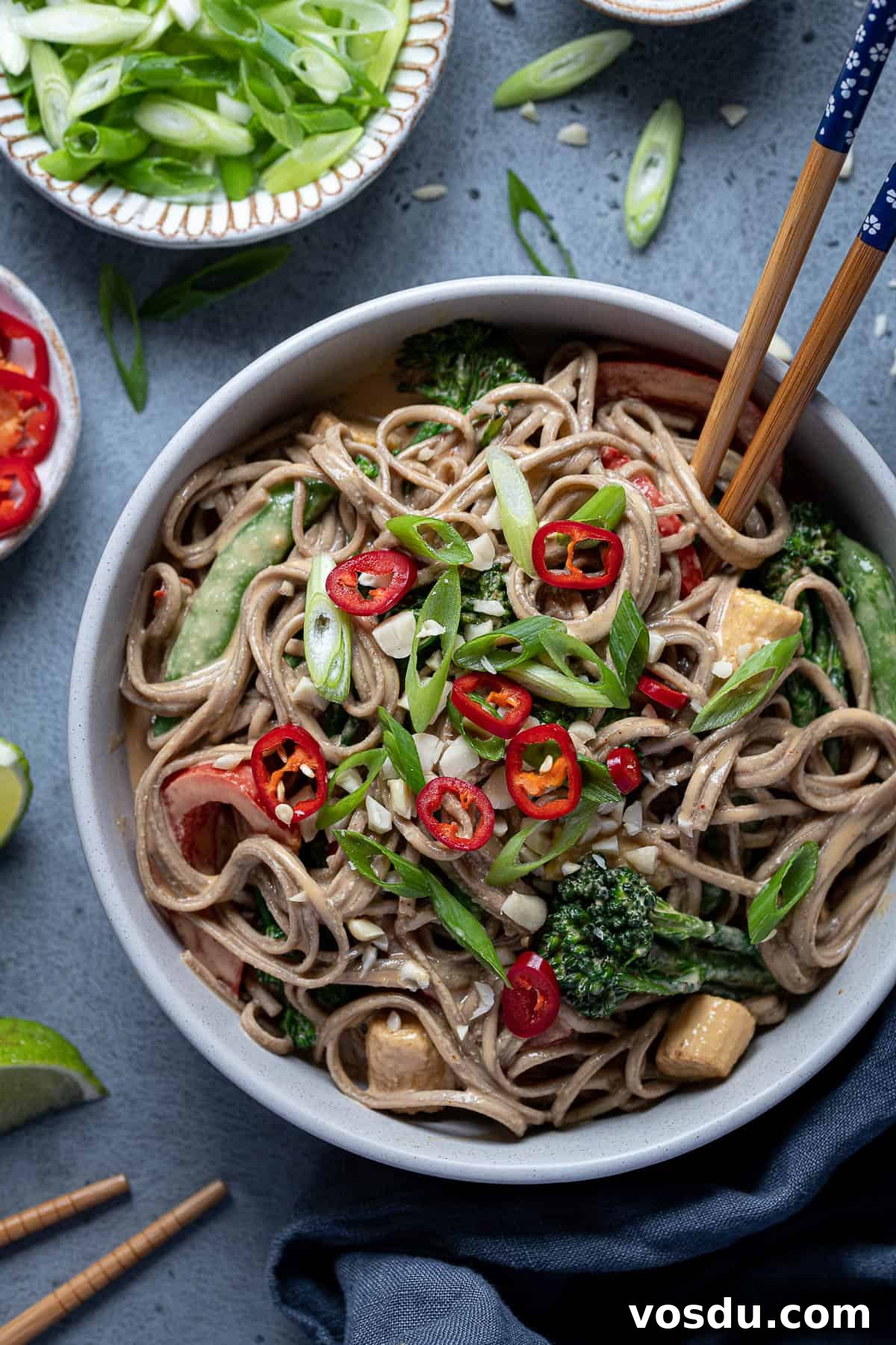 Close up of creamy peanut noodles and vegetables in a bowl with chopsticks, sliced spring onions and chillies.