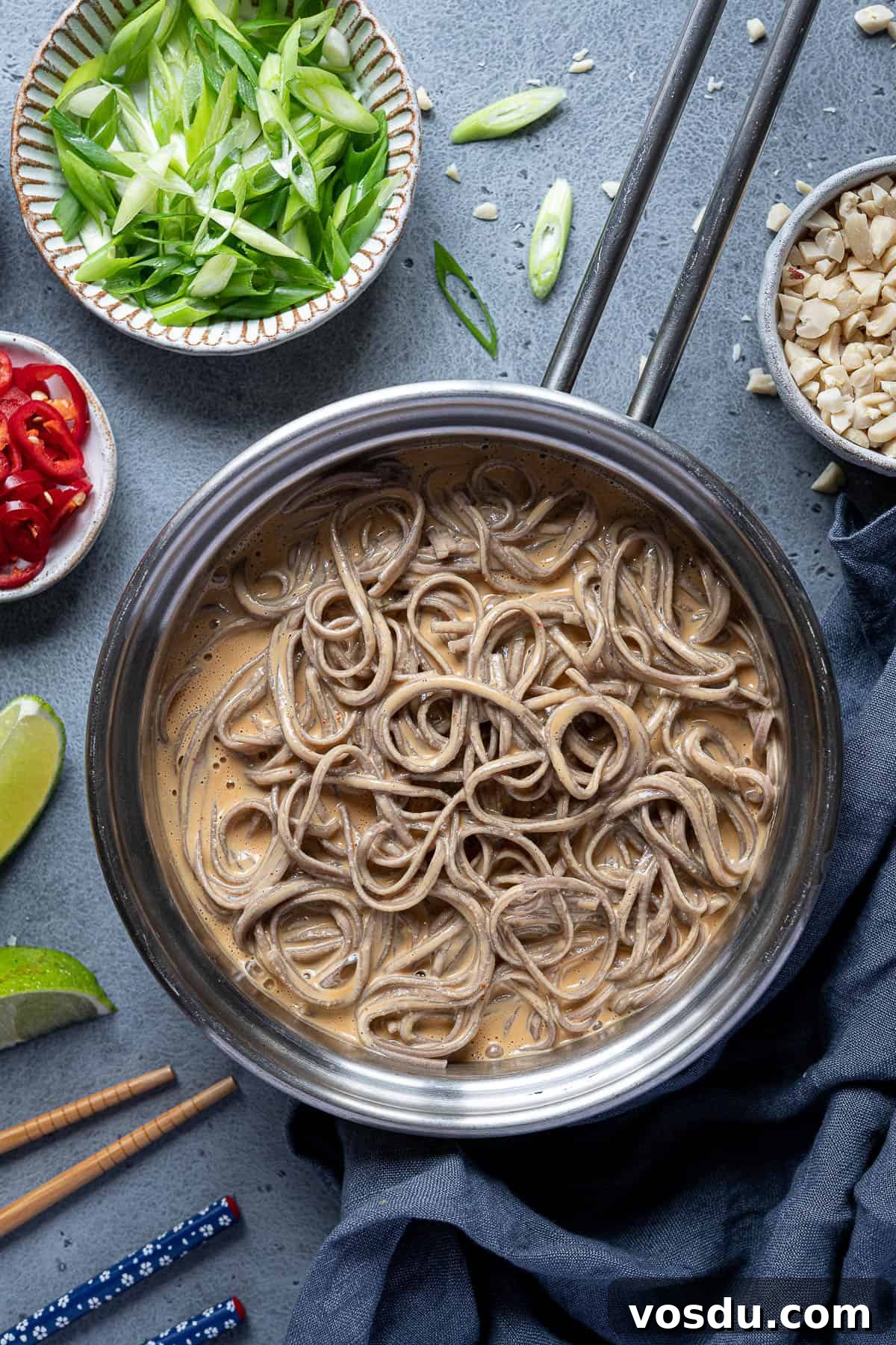 Creamy peanut noodles in a pan with bowls of chopped peanuts, spring onions, chillies and limes.