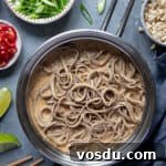 Creamy peanut noodles in a pan with bowls of chopped peanuts, spring onions, chillies and limes.