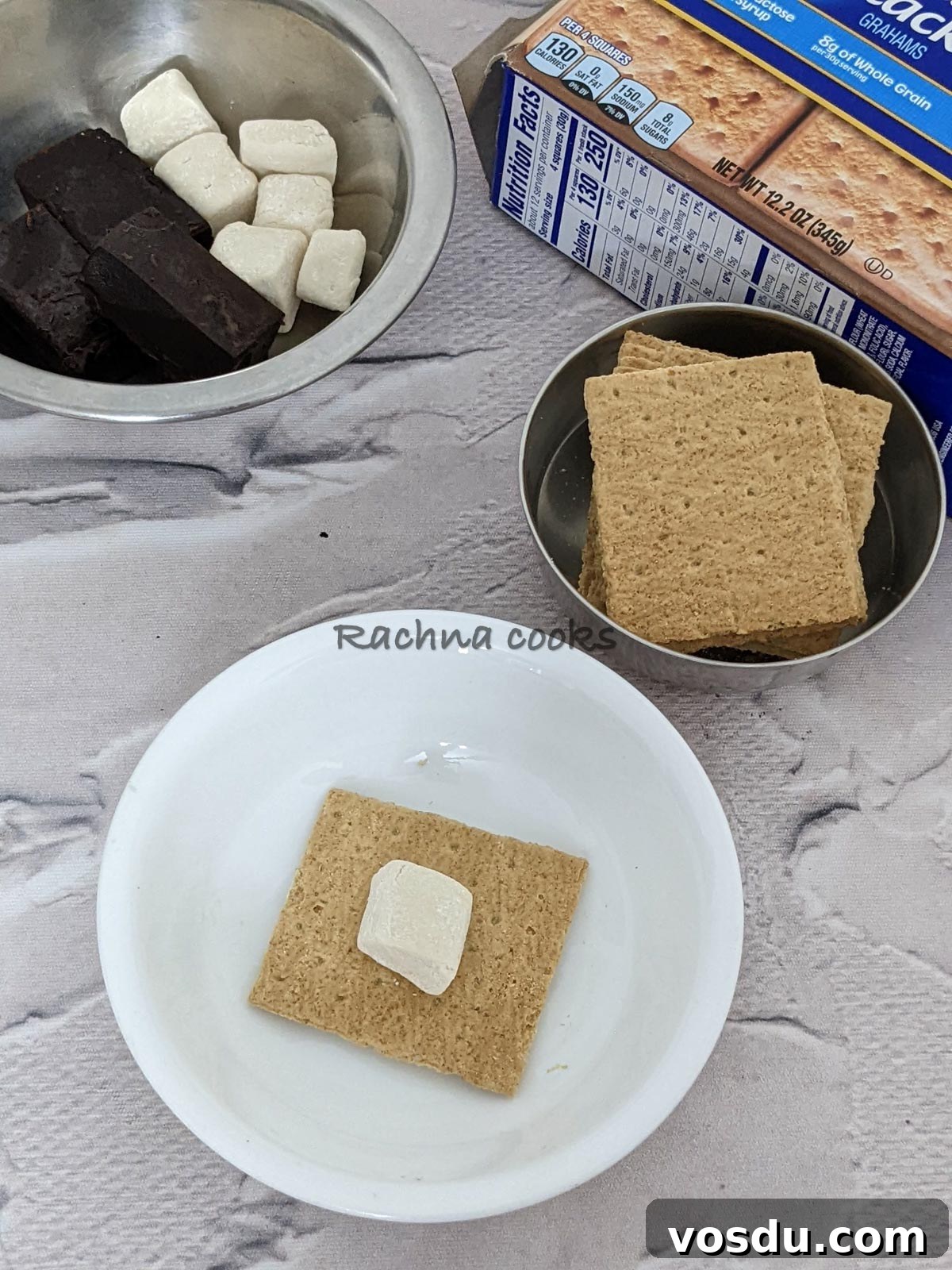 Crispy Air Fryer S'mores Recipe for Quick Desserts 5 Graham's cracker with a pressed marshmallow on top. Bowls of crackers, marshmallows and chocolate bars visible in the background.