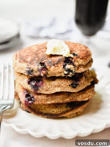 A stack of blueberry oatmeal pancakes have a glass of coffee behind them.
