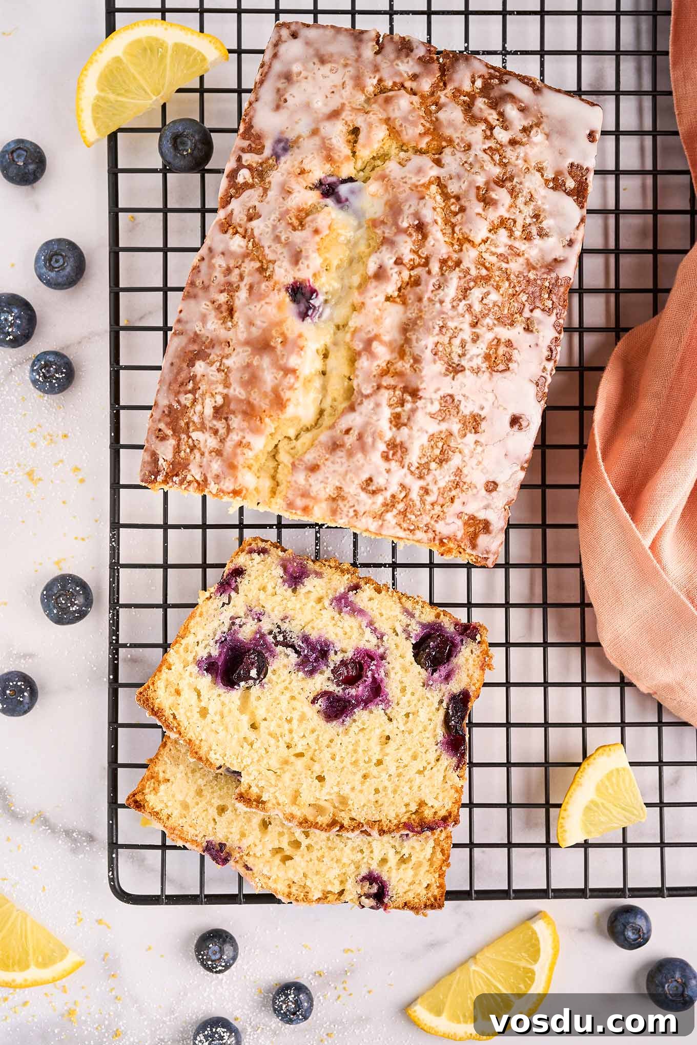 Lemon Blueberry Loaf Cake Recipe with Zesty Glaze 2 Freshly baked lemon blueberry loaf cake on a black wire rack. Slices reveal moist crumb studded with plump blueberries, drizzled with a sweet lemon glaze.