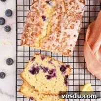 Lemon Blueberry Loaf Cake Recipe with Zesty Glaze 5 Freshly baked lemon blueberry loaf cake on a black wire rack. Slices reveal moist crumb studded with plump blueberries, drizzled with a sweet lemon glaze.