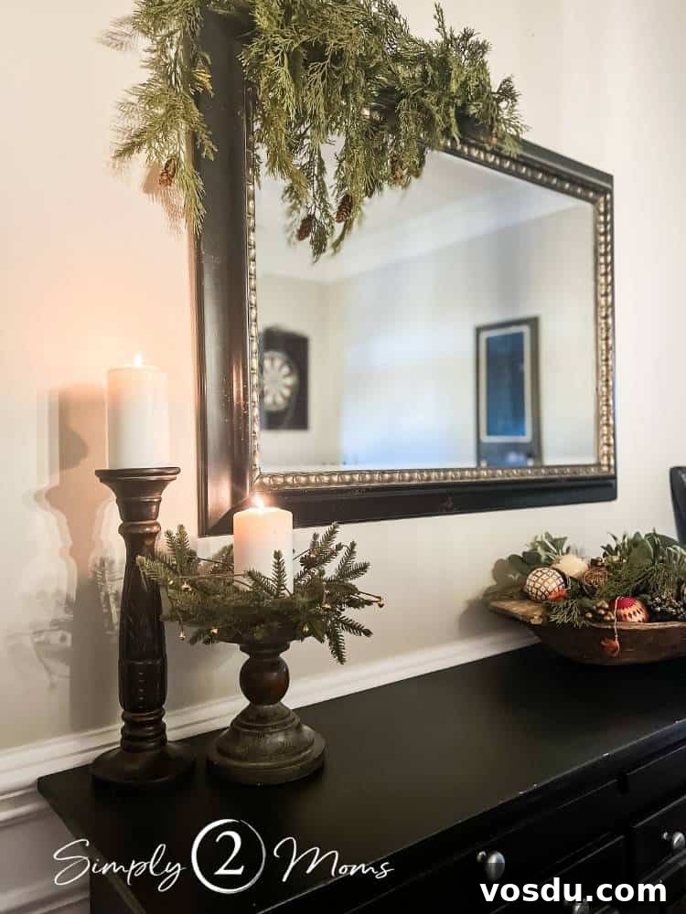 Country Chic Christmas Home Tour That Inspires Holiday Decorating 11 Large rectangular mirror with black and gold frame decorated for Christmas with faux cedar greenery draped over one corner hanging above a black sideboard with wood pillar candle holders and an antique dough bowl filled with ornaments and greenery.