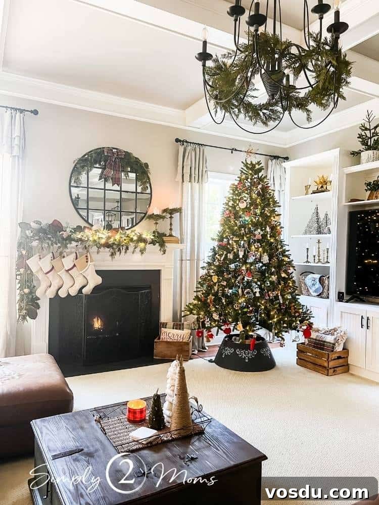 Country Chic Christmas Home Tour That Inspires Holiday Decorating 14 A family room with a gas fireplace with painted marble surround decorated for Christmas with natural greenery garland, custom South House Design stockings, with a large round mirror decorated with a half wreath. A Christmas tree with custom collar is next to the fireplace and an iron chandelier is decorated with more greenery.