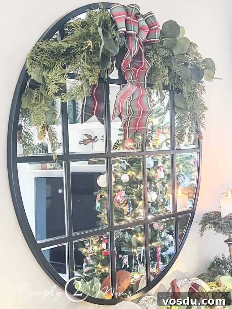 Country Chic Christmas Home Tour That Inspires Holiday Decorating 16 A large round mirror with window pane dividers decorated for Christmas with a half wreath made of faux cedar greenery and eucalyptus and a plaid bow. A Christmas tree is reflected in the mirror.