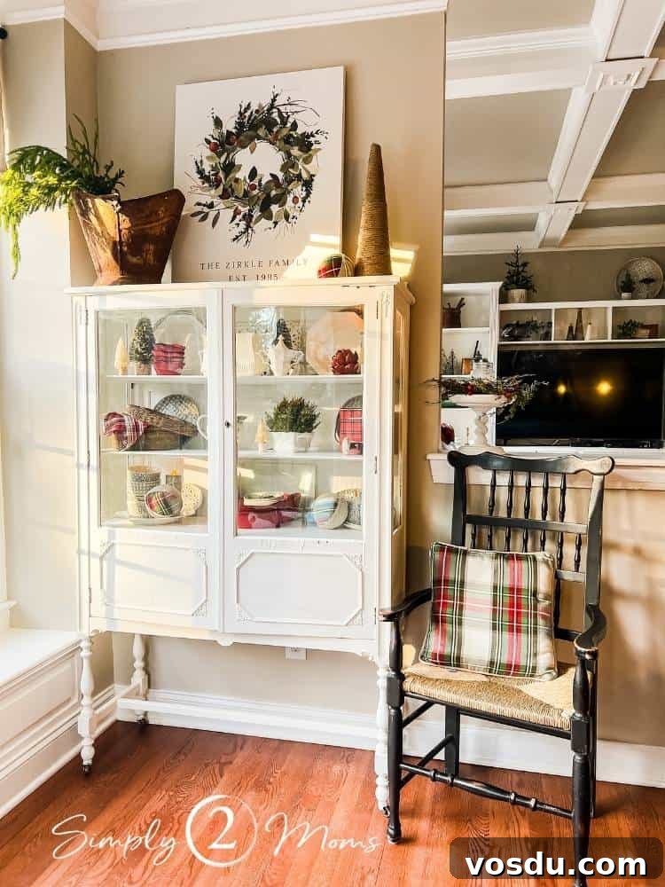 Country Chic Christmas Home Tour That Inspires Holiday Decorating 21 Vintage china cabinet decorated for Christmas with white and red dishes, greenery, and a custom Canvas on Demand canvas. Antique black arm chair with caned seat and Christmas plaid pillow sits next to the hutch.