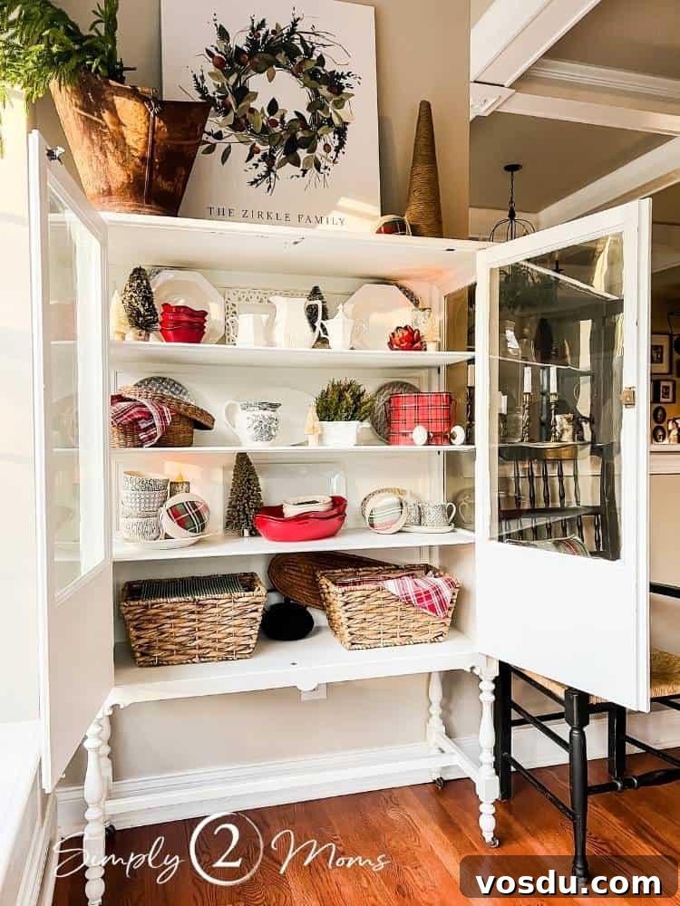 Country Chic Christmas Home Tour That Inspires Holiday Decorating 22 Vintage china cabinet with open glass doors decorated for Christmas with white and red dishes, greenery, and red plaid napkins. On top of the cabinet is an antique coal skuttle filled with greenery, a custom Canvas on Demand canvas, and chunky yarn tree.