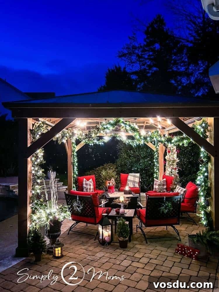 Country Chic Christmas Home Tour That Inspires Holiday Decorating 23 Poolside paver patio with large gazebo decorated for Christmas with lighted garland and string lights. Iron furniture with red cushions sit around a coffee table with a small firepit on top at nighttime.