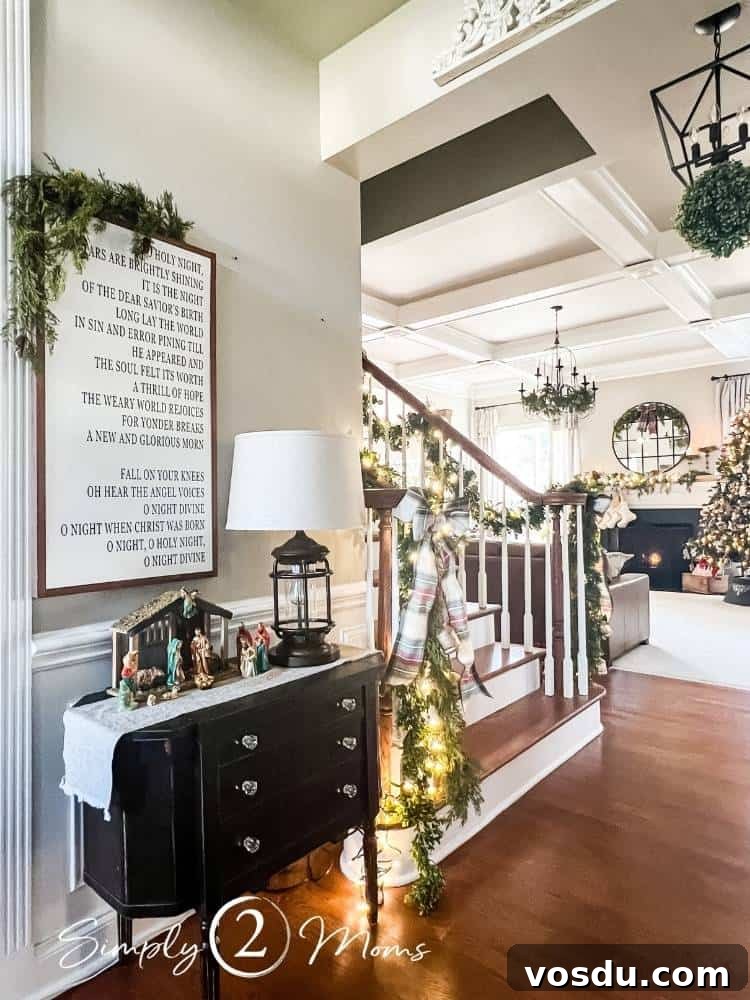 Country Chic Christmas Home Tour That Inspires Holiday Decorating 8 A front entry hallway decorated for Christmas with a nativity scene on a small black antique table. A custom sign from Between You and Me Signs with the lyrics to O Holy Night hangs above the nativity. Beyond the table is a staircase with garland on the banisters and a family room with a fireplace and Christmas tree.
