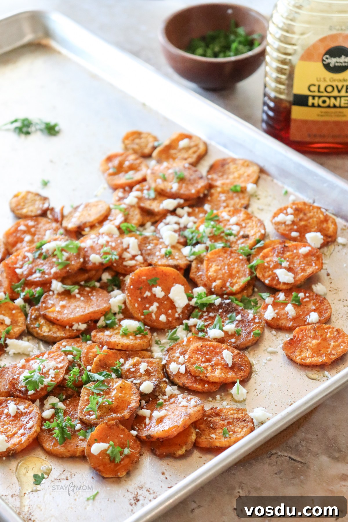 Honey-Roasted Sweet Potato Rounds with Maple Glaze 6 Sheet pan with completed recipe showing sweet potato rounds, roasted, topped with feta and parsley with a bottle of honey in the background.