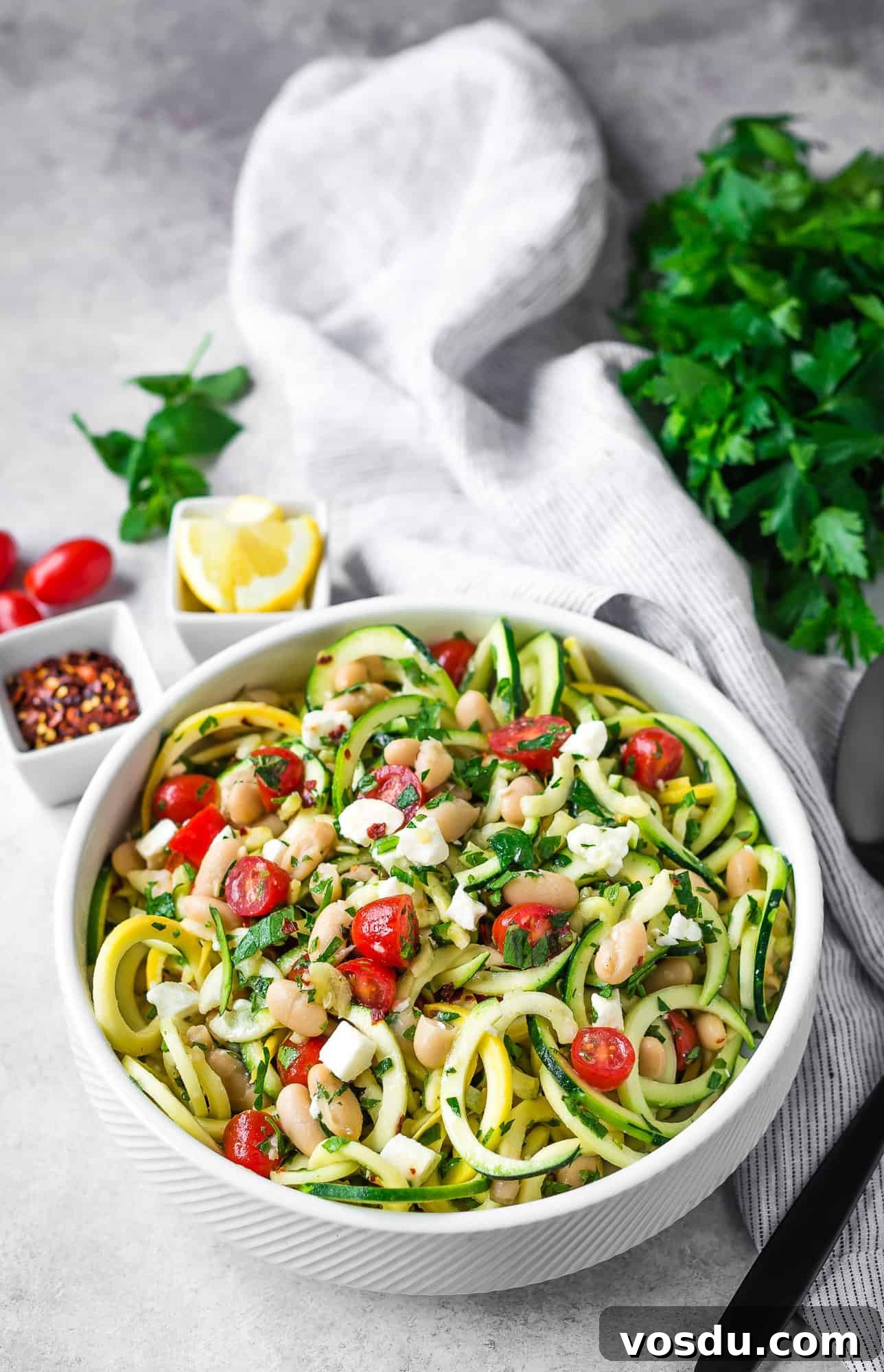 Zucchini Noodle Salad with White Beans, Feta & Lemon Vinaigrette 4 Salad served in a shallow white bowl, additional salad ingredients nearby.