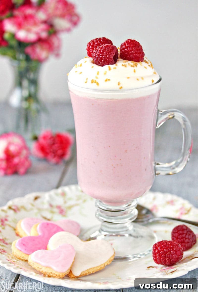 Raspberry White Hot Chocolate is a pretty pink twist on traditional hot chocolate! It's made with white chocolate and real raspberry puree, for a fresh, fruity taste that is perfect year-round! | From SugarHero.com Raspberry White Hot Chocolate - A single cup of hot chocolate with raspberries on top and heart shaped cookies to the side | From SugarHero.com