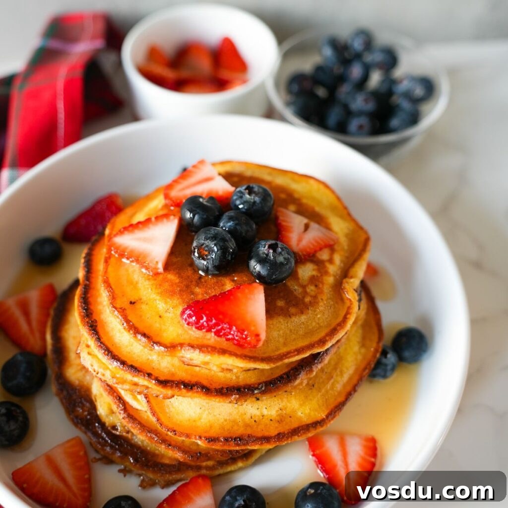 A stack of fluffy lemon ricotta pancakes topped with blueberries, strawberries and drizzled with maple syrup.