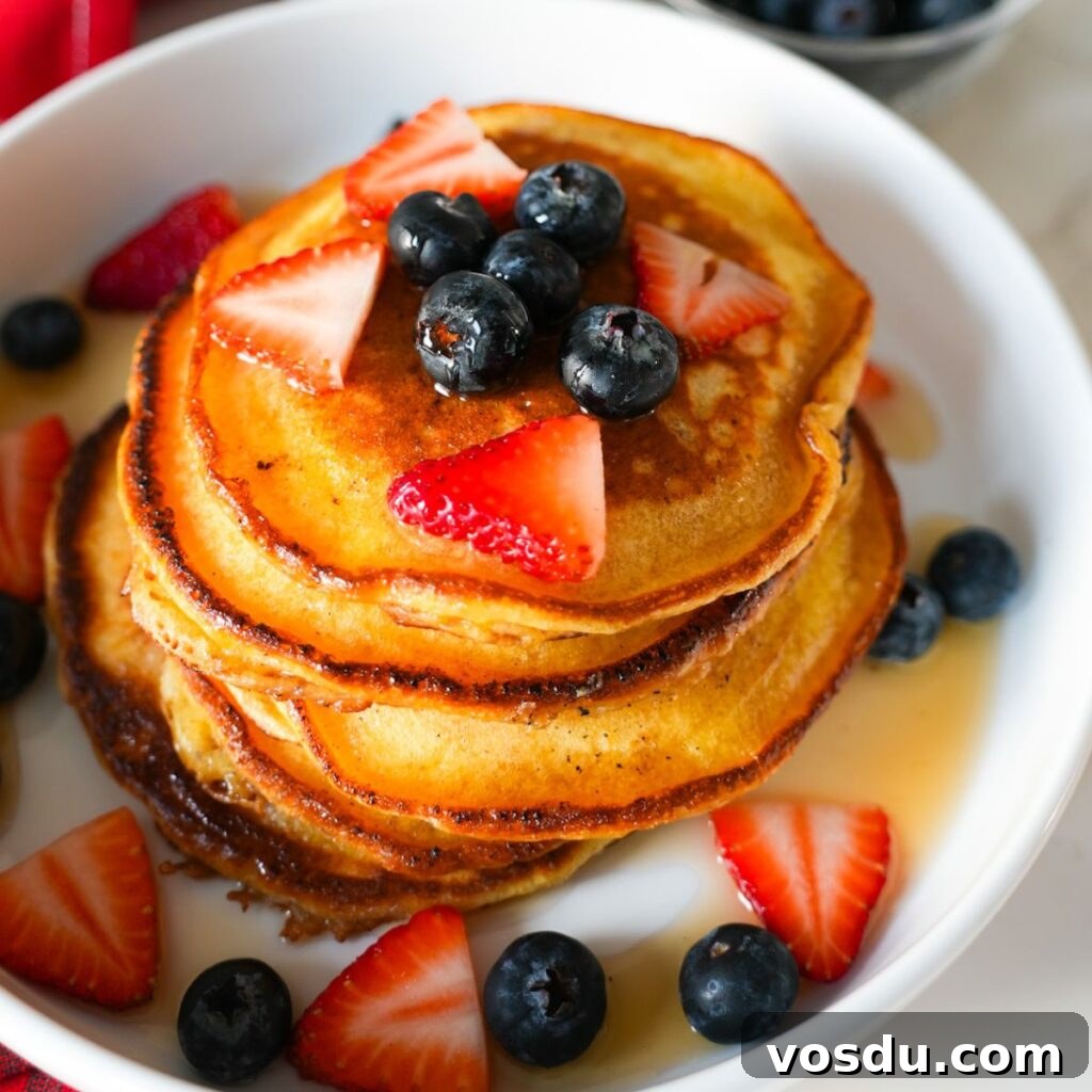 A stack of fluffy lemon ricotta pancakes topped with blueberries, strawberries and drizzled with maple syrup.