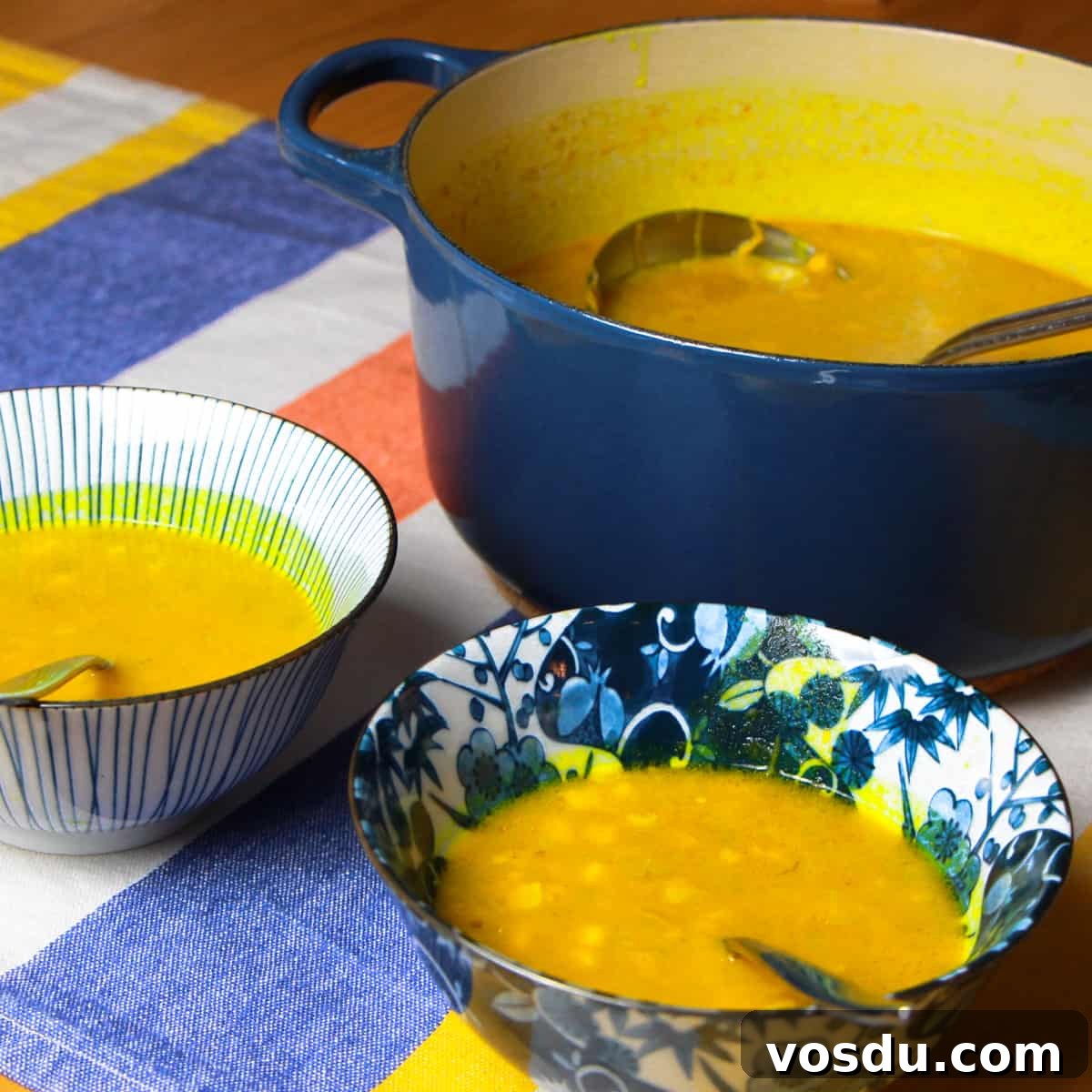 Spiced Pumpkin & Sweetcorn Soup Recipe for Cozy Fall Nights 2 a big navy le creuset pot full of spiced pumpkin and sweetcorn soup with two bowls of soup in the foreground. All on a stripy blue, yellow, orange and white tablecloth on a wooden table.