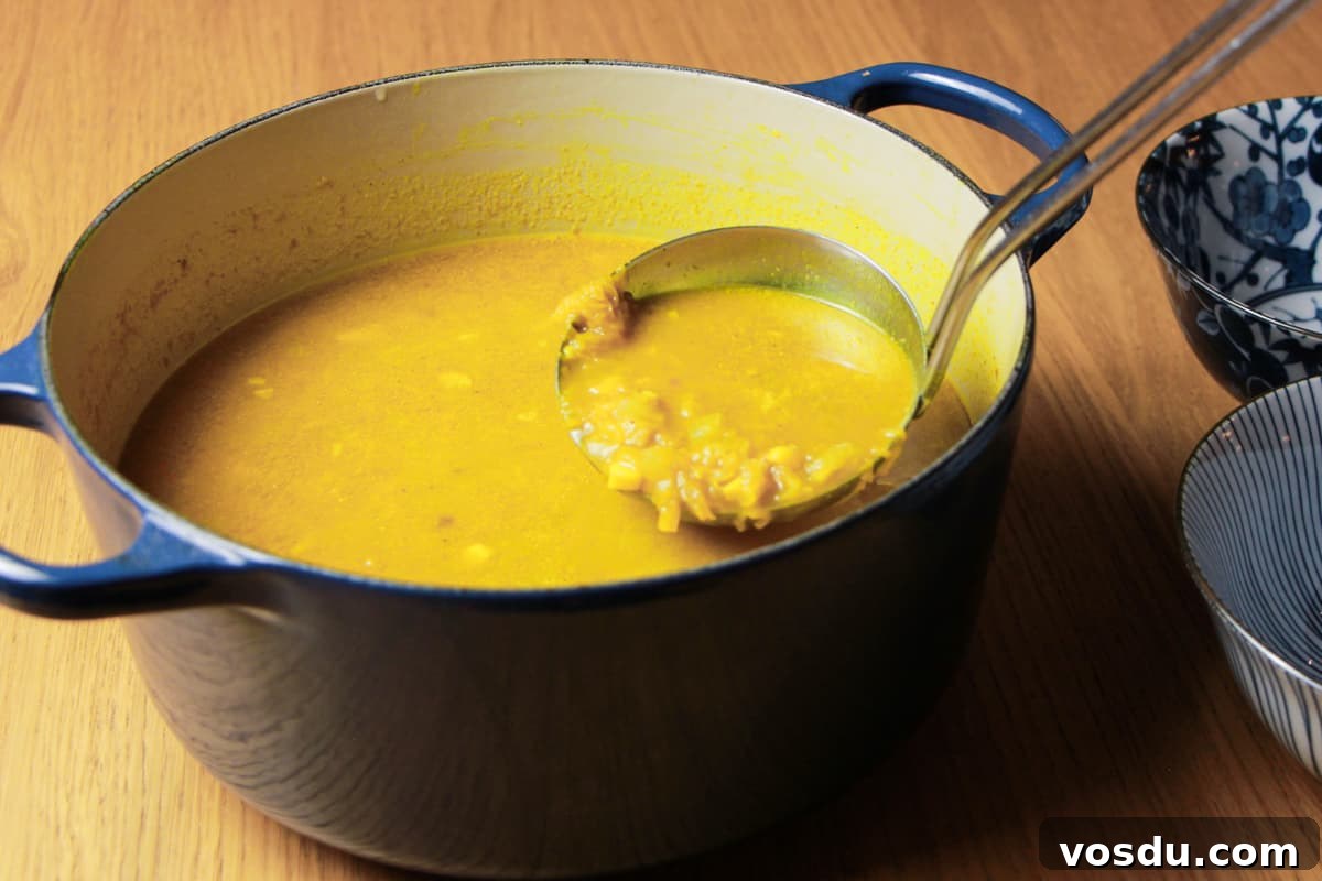 Spiced Pumpkin & Sweetcorn Soup Recipe for Cozy Fall Nights 3 Navy casserole pan full of spiced pumpkin and sweetcorn soup and a ladle taking a scoop out of the pan to go in the two blue and white decorated bowls next to it. All on a wooden table.