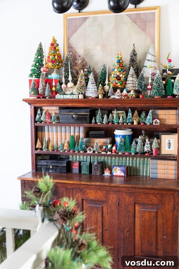 This Year’s Holiday Home Decor: Fresh Ideas to Transform Your Space 13 Dark wood cabinet piled with all sizes of Christmas bottle brush trees and small figures with a painting and spot lights above