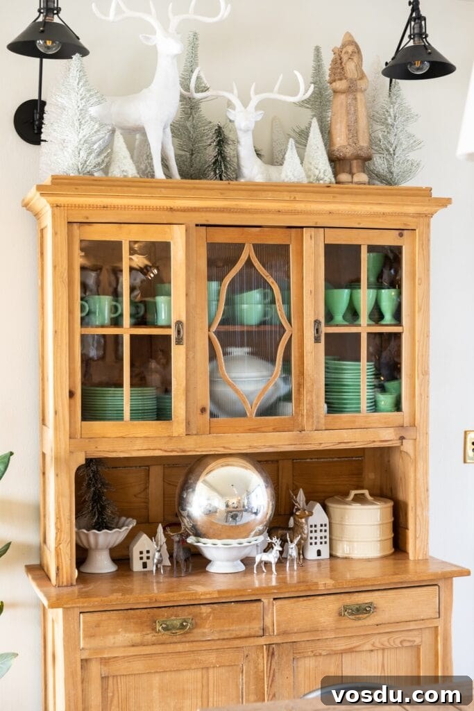 This Year’s Holiday Home Decor: Fresh Ideas to Transform Your Space 18 Light pine colored cabinet filled with green mugs and plates topped with white deer and Christmas trees