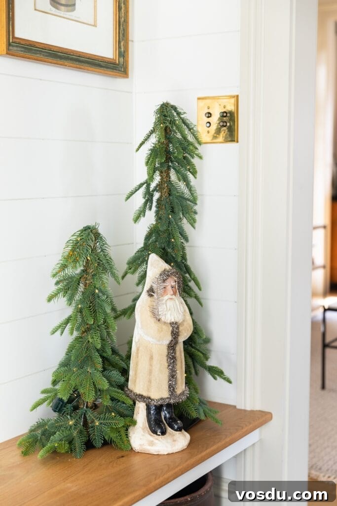 This Year’s Holiday Home Decor: Fresh Ideas to Transform Your Space 4 Small white coated toy Santa standing on wooden bench with two green trees with lights right behind