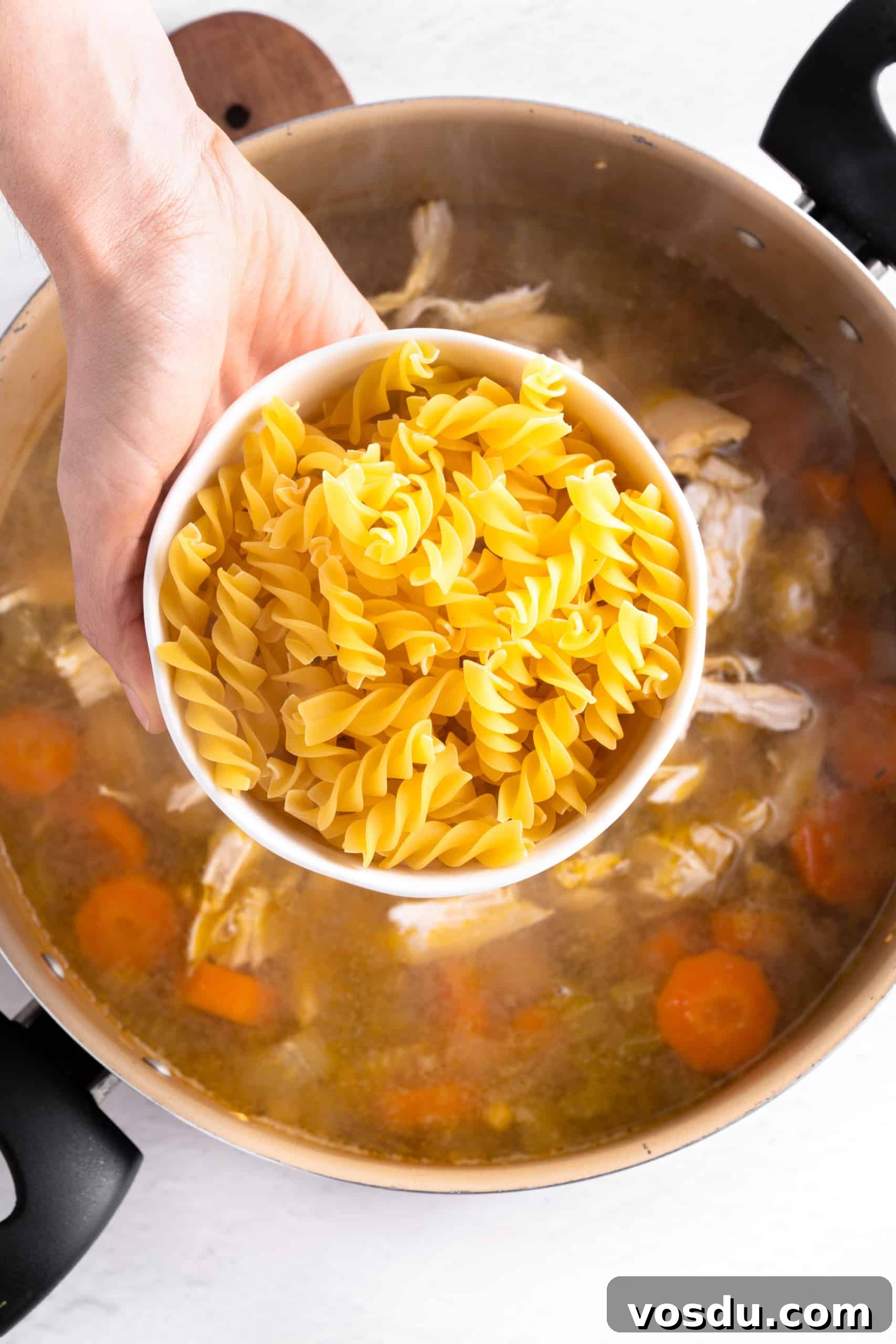 Gluten-Free Chicken Noodle Soup (Dairy-Free) Holding a bowl of fusilli pasta over a pot of soup.