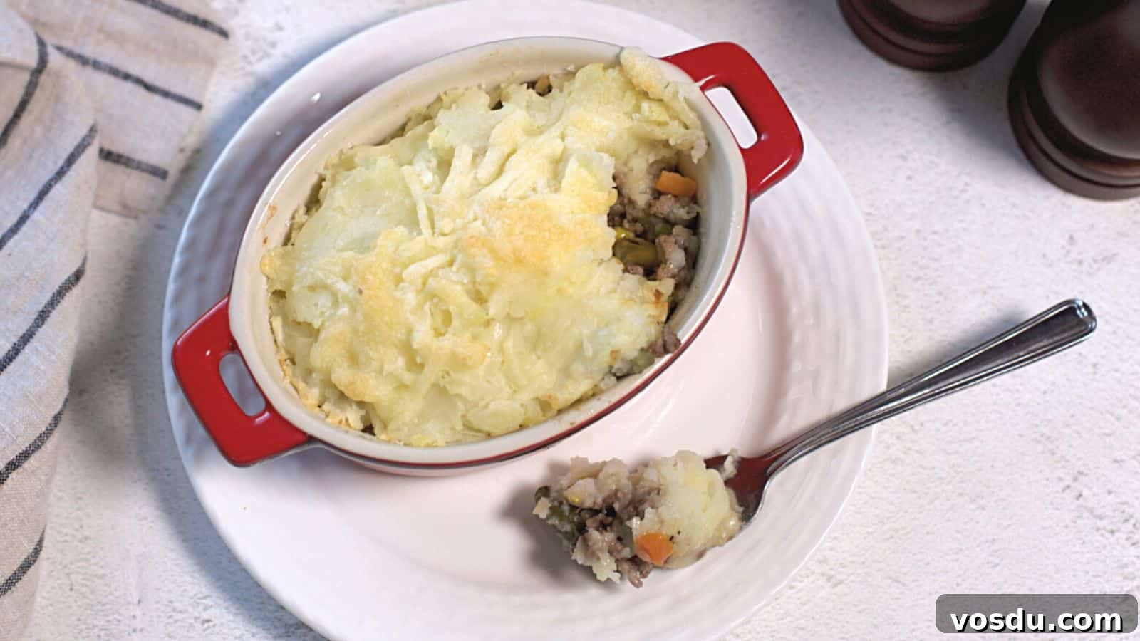 Shepherd's pie in a red ceramic dish with a spoon, showcasing layers of mashed potatoes and minced meat with vegetables on a white tablecloth.