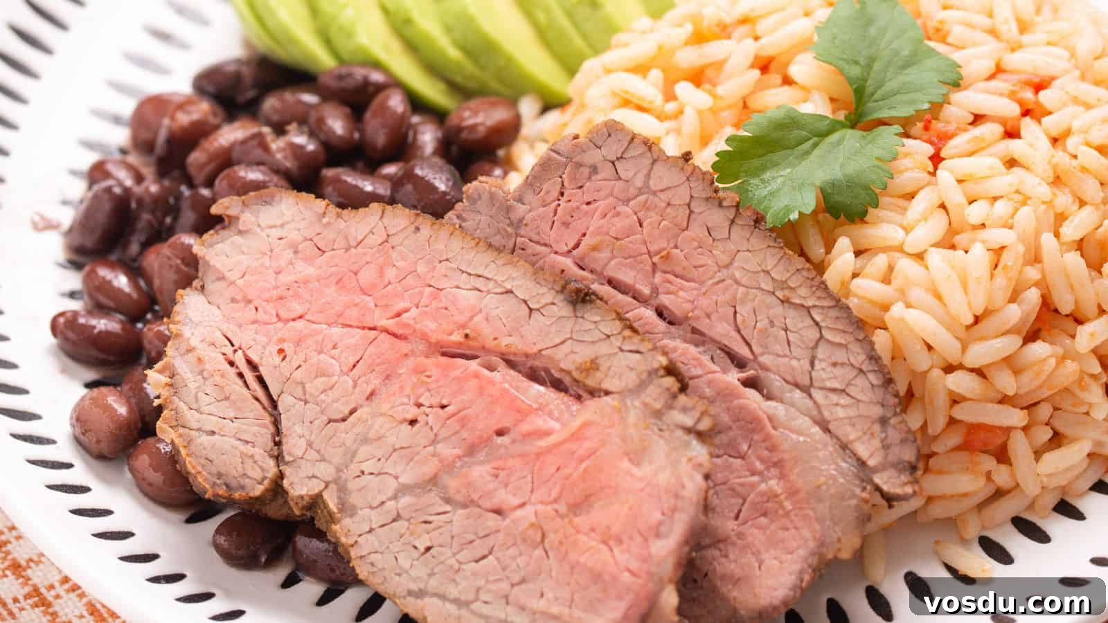 A plate of sliced carne asada with sides of white rice, black beans, and sliced avocado, garnished with a sprig of parsley.