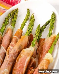 Chilled Asparagus and Tomato Salad with Lemon Vinaigrette 7 A plate of succulent, prosciutto-wrapped asparagus spears, neatly arranged on a white surface with a vibrant cloth in the background.