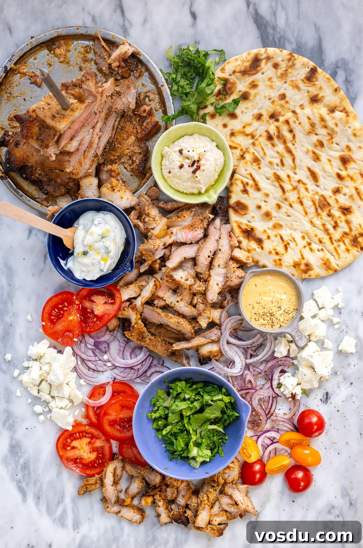 Crispy Air Fryer Pork Gyros Recipe for Quick Weeknight Meals 3 Deconstructed Air Fryer Pork Gyros ingredients on a marble surface: grilled meat, pita bread, tzatziki, hummus, mustard sauce, sliced tomatoes, red onions, feta cheese, leafy greens, chopped herbs, and cherry tomatoes.