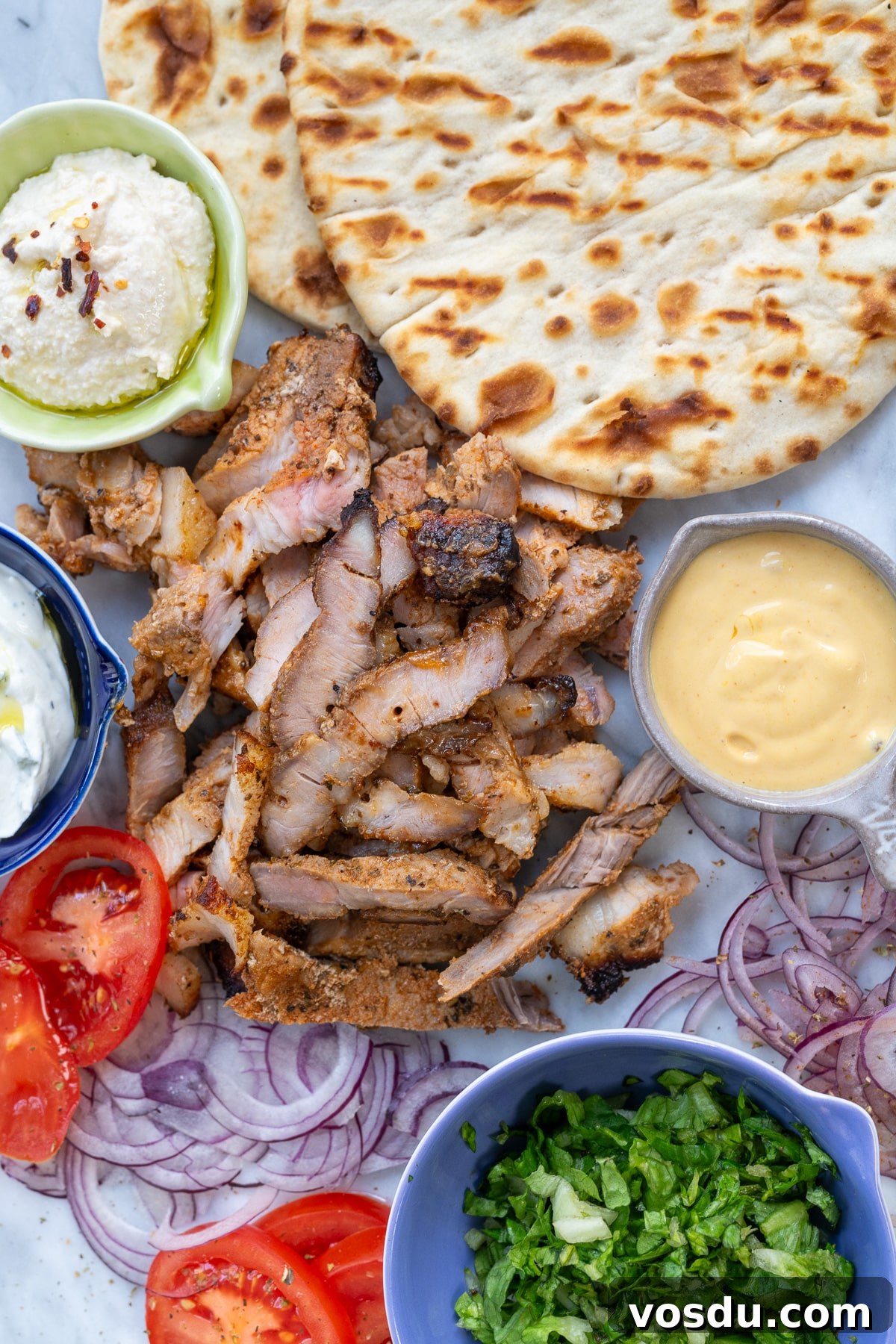 Crispy Air Fryer Pork Gyros Recipe for Quick Weeknight Meals 4 A platter featuring Pork Gyros with sliced grilled meat, two pieces of flatbread, sliced red onions, tomato, chopped lettuce, a bowl of yellow sauce, tzatziki, and a bowl of whipped feta cheese—an authentic Greek recipe delight.