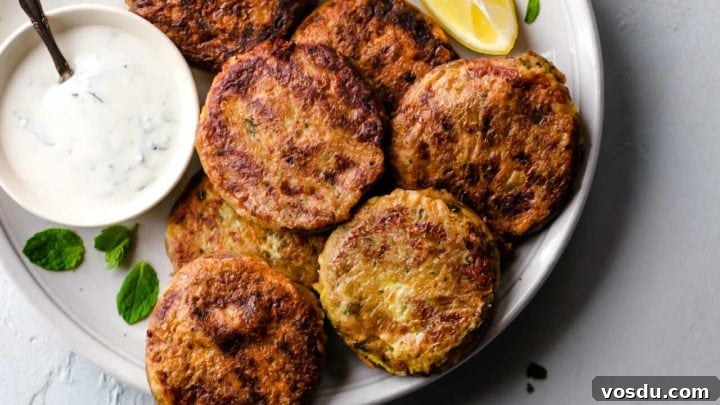 A white plate with Shami Kebab alongside a bowl of raita, lemon wedge and garnished with mint leaves.