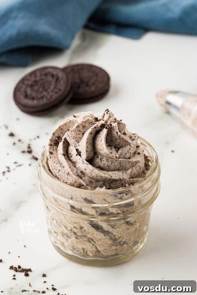 Cookies and Cream Frosting Recipe piped into a small glass jar with Oreo cookies and a blue kitchen towel in the background