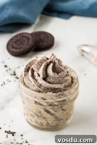 Cookies and Cream Frosting Recipe piped into a small glass jar with Oreo cookies and a blue kitchen towel in the background