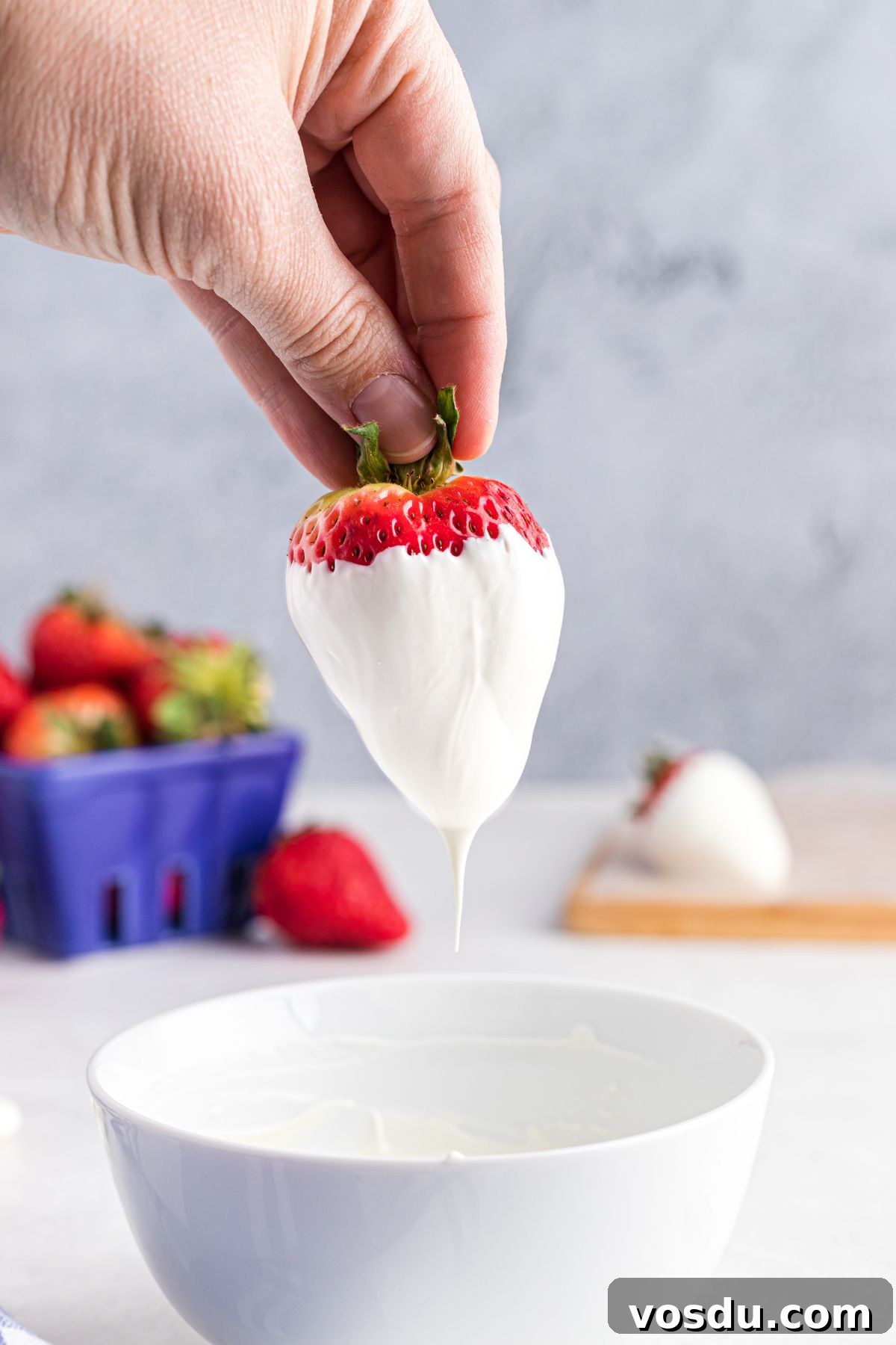 A hand dipping a strawberry in melted white candy coating, in a small white bowl.