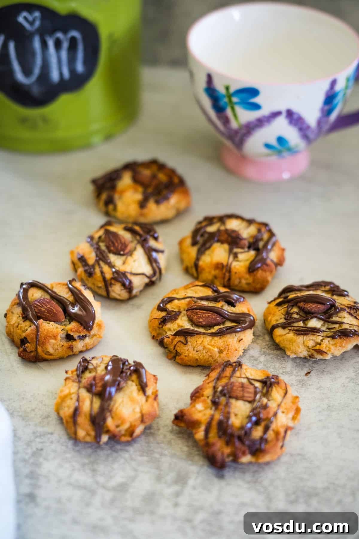 Keto Almond Joy Cookies Recipe — Low-Carb Coconut Chocolate Cookies 2 Chocolate-drizzled cookies with almonds are arranged on a table next to a decorative cup and a green container labeled "YUM.