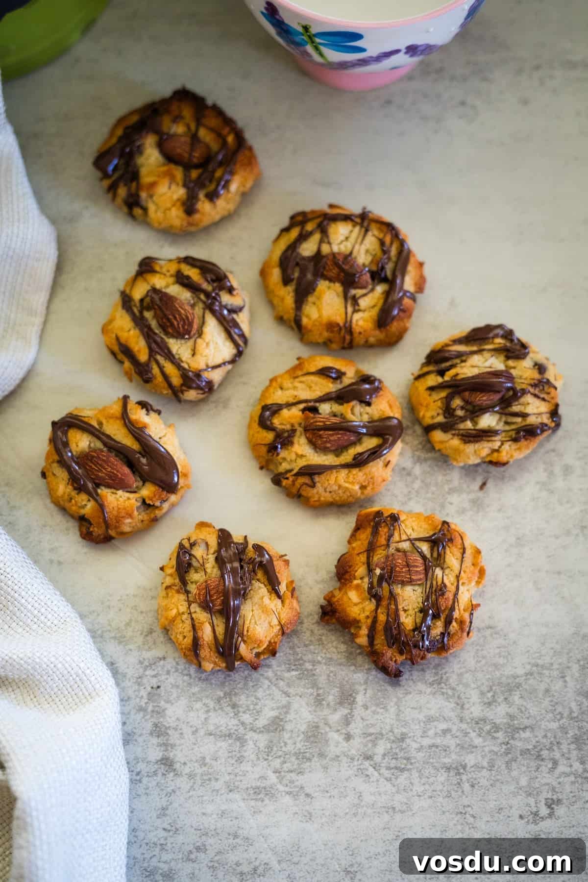 Keto Almond Joy Cookies Recipe — Low-Carb Coconut Chocolate Cookies 11 A group of keto almond joy cookies drizzled with chocolate are arranged on a gray surface. A white cloth and a floral cup are partially visible nearby.