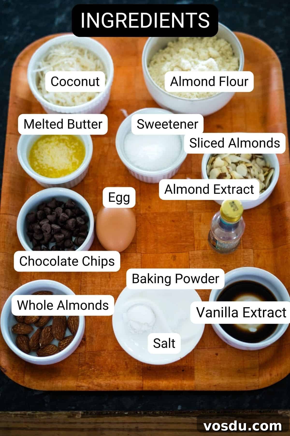 Keto Almond Joy Cookies Recipe — Low-Carb Coconut Chocolate Cookies 3 Wooden tray with labeled ingredients: coconut, almond flour, melted butter, sweetener, sliced almonds, egg, almond extract, chocolate chips, whole almonds, baking powder, salt, and vanilla extract.