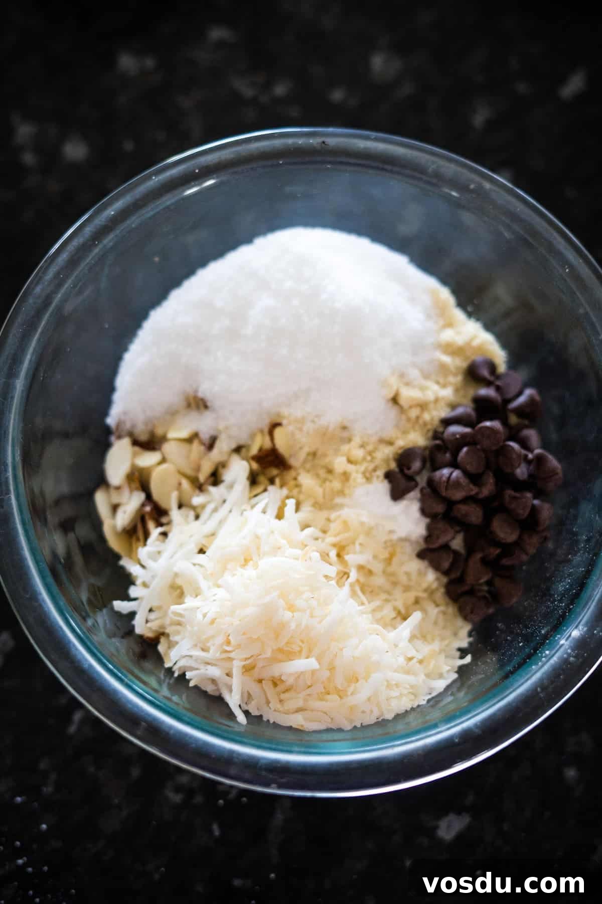 Keto Almond Joy Cookies Recipe — Low-Carb Coconut Chocolate Cookies 4 A glass bowl containing shredded coconut, chocolate chips, sliced almonds, and sugar, placed on a dark surface.