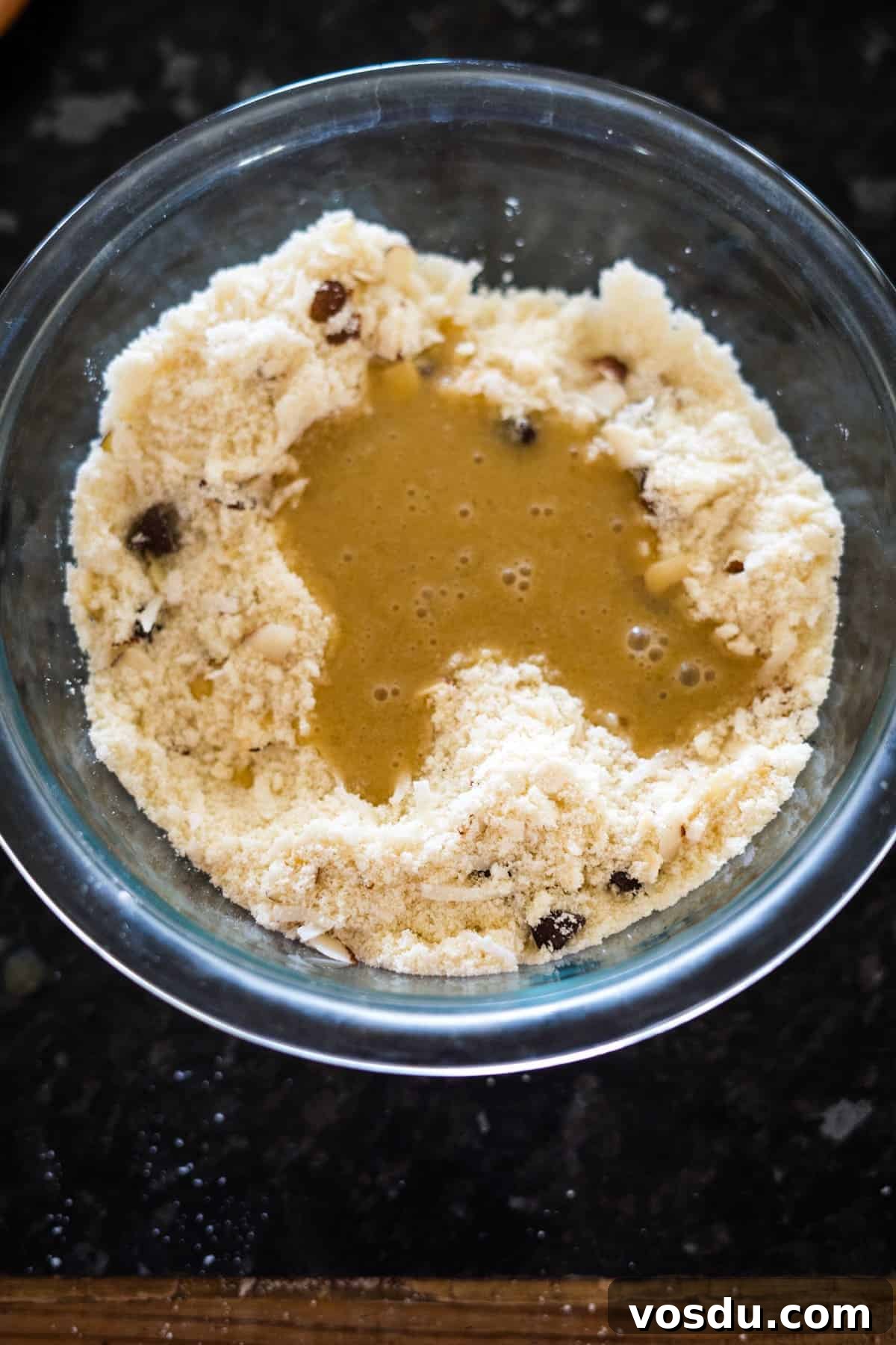 Keto Almond Joy Cookies Recipe — Low-Carb Coconut Chocolate Cookies 6 A glass bowl with flour mixture and raisins, with a pool of liquid in the center, on a dark countertop.