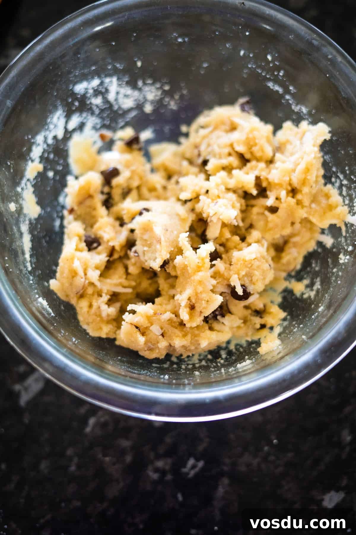 Keto Almond Joy Cookies Recipe — Low-Carb Coconut Chocolate Cookies 7 A glass bowl containing chunky cookie dough with visible chocolate chips on a dark countertop.